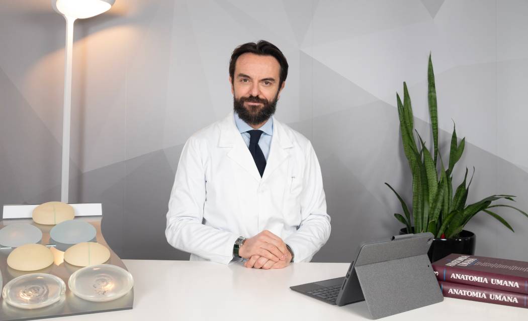 Foto del Dottor Valerio Badiali, chirurgo estetico presso IEI - Istituto Estetico Italiano, in camice bianco seduto a una scrivania con alla sua sinistra delle protesi utilizzate per gli interventi di mastoplastica e alla sua destra un tablet e dei libri di anatomia umana.